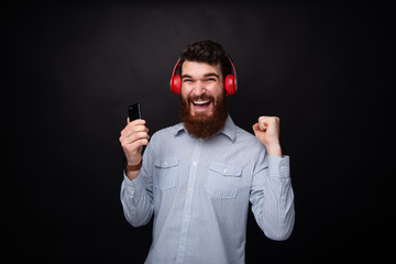Photo of cheerful bearded young man, celebrating with arms up, while using a mobile phone and headphoneas