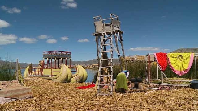 Medium low-angle still shot of Uros village compound with a ladder leaning on lookout tower, coloured traditional skirts, reed boat, and a woman in front of a  cooking stove, Uros Village, Peru