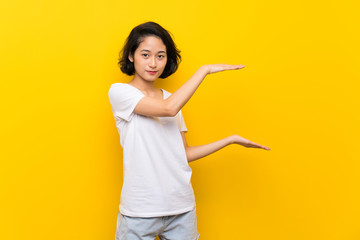 Asian young woman over isolated yellow wall holding copyspace to insert an ad