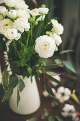 Spring white buttercup flowers in white enamel jug on wooden vintage chair with curtain behind,...