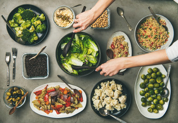 Vegan dinner table setting. Healthy vegetarian dishes in plates on table. Flat-lay of vegetables, legumes, beans, olives, sprouts, hummus, couscous and woman hands taking fresh salad from bowl, top
