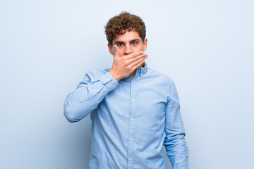 Blonde man over blue wall covering mouth with hands