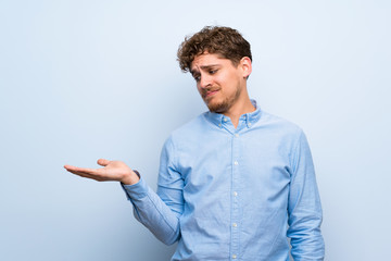 Blonde man over blue wall holding copyspace with doubts