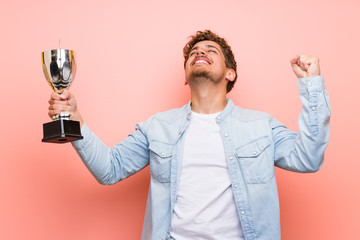 Blonde man over pink wall holding a trophy