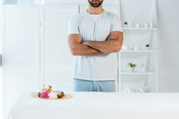cropped view of man standing with crossed arms near tray with bottles of oil, sea salt and flowers