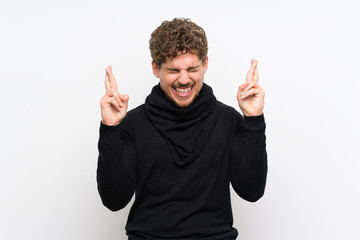 Blonde man over isolated white wall with fingers crossing