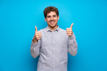 Blonde man over blue wall with thumbs up gesture and smiling