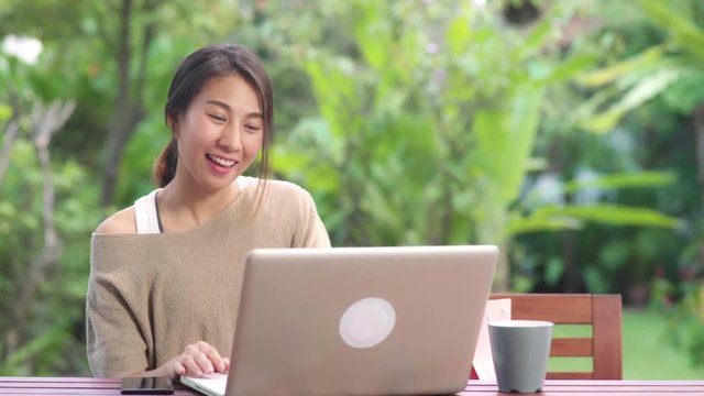 Asian woman using laptop video conference with friends, female relax feeling happy showing shopping bags sitting on table in the garden in morning. Lifestyle women relax at home concept.
