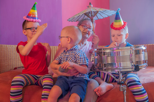 Elderly woman in pink T-shirt and glasses holding megaphone. Next to grandmother, grandchildren in festive clothes in clown hats and patch on nose are playing with drum. Gray speak louder in hand