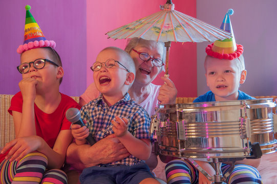 Elderly woman in pink T-shirt and glasses holding megaphone. Next to grandmother, grandchildren in festive clothes in clown hats and patch on nose are playing with drum. Gray speak louder in hand