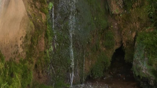 A tilting close up shot of water running down a moldy rock as it drips into a small pond with the sun shing brightly on a warm afternoon