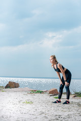 tired sportive young woman standing on beach near sea