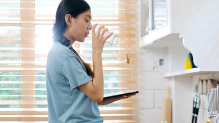 Young beautiful asian woman drinking water while holding digital tablet and standing by window in kitchen background, peolpe and healthy lifestyles