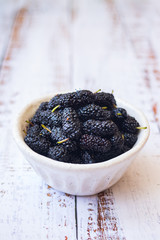 Sweet ripe black mulberry berries in a bowl. On wooden white table.