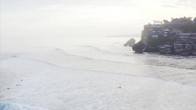 Flying backwards above Uluwatu spot and surfers waving on Indian ocean waves. On the background is cliff with houses, cfes, resorts, etc. Aerial view. Bali, Indonesia