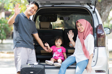 young muslim family , transport, leisure, road trip and people concept - happy man, woman and little girl standing and sitting  on trunk of hatchback car and waving hand outdoors