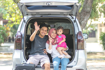 young muslim family , transport, leisure, road trip and people concept - happy man, woman and little girl sitting on trunk of hatchback car and waving hand outdoors