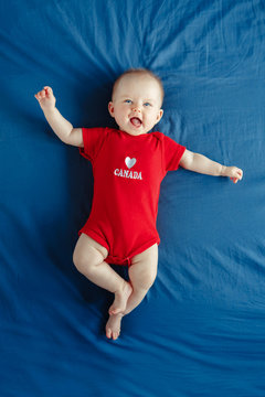 White Caucasian smiling baby boy girl with blue eyes lying on bed at home on Canada Day. Newborn infant child in red onesie romper celebrating national holiday July 1. View from top above