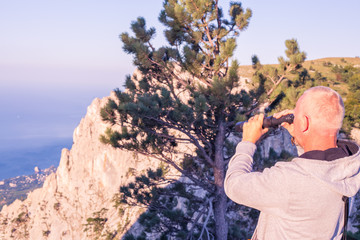 a man with blond hair stands on the edge of Ai-Petri mountain in the Crimea against the background of the sea and rocks and looks through a telescope