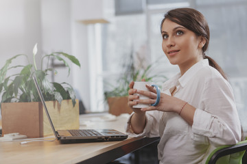 Young beautiful businesswoman looking away thoughtfully, enjoying her coffee at the office, copy space. Charming female entrepreneur having morning coffee at work. Achievement, startup concept