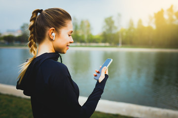 Young smiling girl making sport and running in the park using her phone to listen the music with...