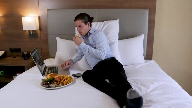 Business Man On Hotel Bed Eats Food And Works On Laptop. A businessman laying on his hotel bed works on his laptop while eating a fry.