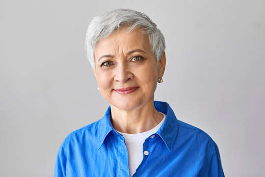 Age and beauty concept. Charming positive mature European female with short gray hair and wrinkles posing isolated, looking at camera with confident smile, wearing stylish blue shirt. Studio image