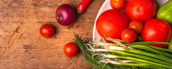 Fresh tomatoes, cherry tomatoes, green, chili peppers, onions, cucumbers, garlic and herbs in a plate on a rustic wooden table - panoramic culinary background, copy space, healthy eating concept