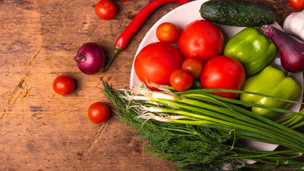 Fresh tomatoes, cherry, green peppers, chili peppers, onions, cucumbers, garlic and herbs in a plate on a rustic wooden table - culinary background, copy space, rural style, healthy eating concept
