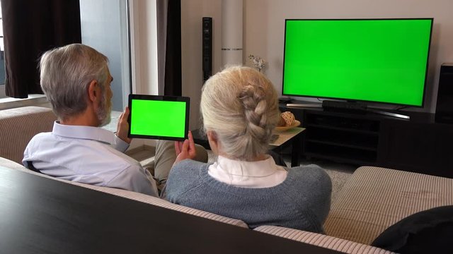 An elderly couple sits on a couch in a living room, watches TV with a green screen and looks at a tablet with a green screen