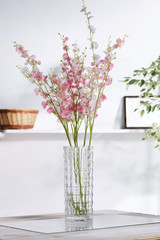 Shot of interior design. There is a floral composition in the middle with whitey-pink orchid twigs in a waterglass. On the blurred background there is a wall shelf with photo frame and woven basket.