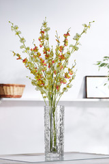 Shot of interior design. In the middle there is a floral composition with orange and green orchid twigs in a vase. On the blurred background there is a wall shelf with photo frame and woven basket.