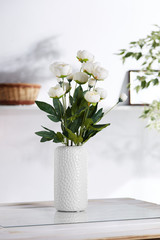 Shot of interior design. The floral composition is in the middle, with bunch of white peonies in a white porcelain vase. On blurred background there is wall shelf with photo frame and woven basket.