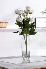 Shot of interior design. The floral composition is in middle, with bunch of white peonies in transparent oval waterglass. On blurred background there is wall shelf with photo frame and woven basket.
