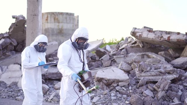 two scientists in protective suits and masks and a personal ionizing radiation dosimeter , walk measure radiation on the background of the ruins of a man-made disaster