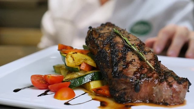 plated steak with chef in background slow motion. Chef stands behind a plated dish in kitchen ready to be served in restaurant. The camera slowly moves around as the chef places hand behind the steak