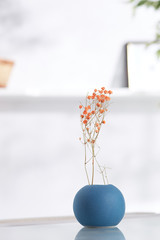 Shot of interior design. There is floral composition in the middle with orange immortelle twigs in a sphere-shaped vase with matte surface. On blurred background there is wall shelf with photo frame.