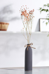 Shot of interior design. There is floral composition in middle with immortelle twigs in bottle-shaped vase adorned with a packthread bow. On blurred background there is wall shelf with a photo frame.