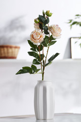 Closeup shot of interior design. There is floral composition in the middle with light brown roses in a snowy bisque vase with ribbed edges. On blurred background there is wall shelf with photo frame.