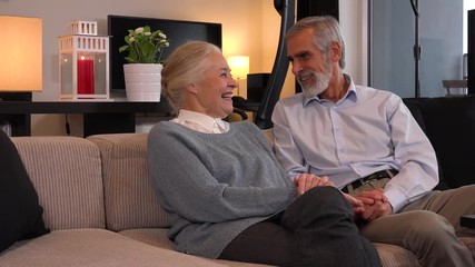 An elderly couple sits on a couch in an apartment, laughs and hugs