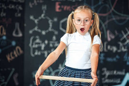 Little blonde girl with a surprised face with a ruler in her hands Pupil girl with big rulers against chalkboard with school formulas. back to school. Schoolgirl learning geometry. math lesson.