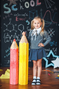 Back to school! A little blonde girl in school uniform stands with two very large pencils on the floore against chalkboard with school formulas at school. The kid is studying in the classroom.