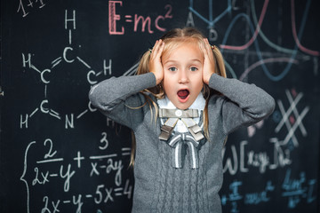 Little girl blonde in school uniform holding hands on her head., open his mouth against chalkboard with school formulas. Complex school program, does not match the age of the child.