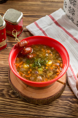 Homemade soup with beans, vegetables and sausages in red bowl on wooden table
