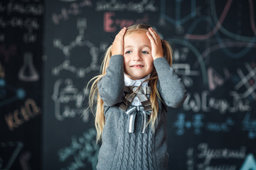 Portrait of little blond girl, grabbing her head. chalkboard with school formulas at the background, concept photo.