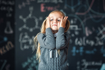 Portrait of little blond girl, grabbing her head. chalkboard with school formulas at the background, concept photo
