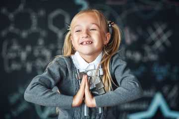 Back to school! A little girl stands with with her hands folded on her chestagainst chalkboard with school formulas at school. The kid is studying in the classroom.