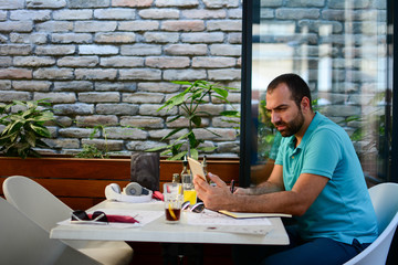 Young man drinking coffee in cafe and using tablet computer, laptop, mobile. Side view.