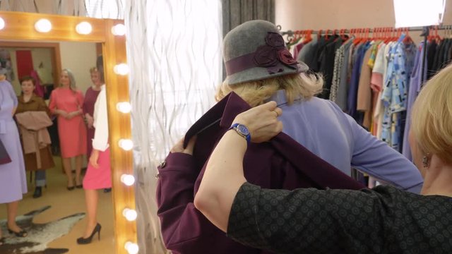 Fashionable senior woman in hat is trying purple coat in clothes store and looking at mirror. Sales consultants help her with the choice.