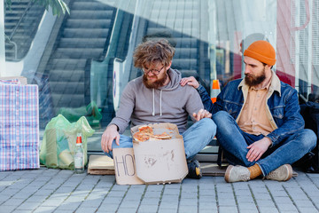 Homeless beggar and young handsome beard man listening to his story sitting together on the ground outdoors. Concept of human understanding